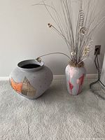 Two pottery vases on carpet, one round gray vase with textured orange and metallic design, and one tall white vase with red streaks and paper decorations