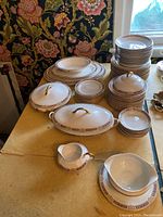 Sixty pieces of porcelain dinnerware including plates, bowls, covered serving dishes, platters, and creamers displayed on a table with decorative floral wallpaper in the background.