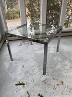 Square chrome and glass table from a low angle showing the entire table and the carpet beneath it, highlighting wear on chrome legs.