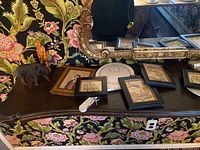 Photo showing carved wood ox, black carved wood bird, Lenox porcelain dish, several framed artworks, and wooden bird figurine on a dark wood table with ornate mirror and floral wallpaper background