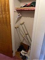 Closet interior showing a wooden door, two leaning aluminum crutches with beige padding, a green cap on top shelf, and other hats partially visible on shelf.