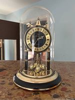 Front view of Elgin American quartz clock inside glass dome showing black and gold dial with roman numerals, brass pendulum with four globes, black column supports and circular black base with gold trim.