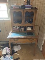 French Provincial style desk with hutch on top, cluttered with papers and items. Desk features slide out leather inset writing surface, visible wear and scratches on wood and leather.