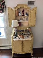 Full view of French Provincial secretary desk with open upper cabinet displaying porcelain figurines, books, lamp on the surface, and assorted decorative items.