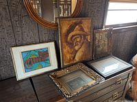 Group image showing 4 framed art pieces leaning on a table against wood paneled wall, and one empty frame resting flat with postcard inside.