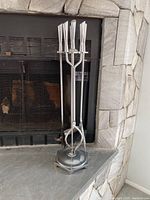 Full view of four metal fireplace tools with mid-century modern style handles held in hexagonal holder in front of stone fireplace.