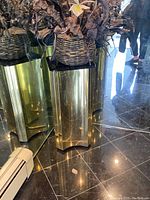 Full view of reflective metal pedestal placed on dark tiled floor with a basket of artificial flowers on top.
