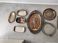 10 silverplate items arranged on floor, showing variety of trays and dishes in different sizes and shapes, some with decorative edges and handles, exhibiting tarnish and patina.