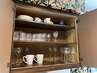 Inside wooden cabinet showing three shelves with items: top shelf stacked dishes and porcelain Coalport teacups and saucers, middle shelf clear glass tumblers and various glasses including one cut glass wine glass, bottom shelf small wine glasses and white ceramic mugs.