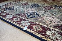 Close-up photo of the corner of a large area rug showing patchwork squares with floral designs in burgundy, blue, and cream colors with a burgundy border