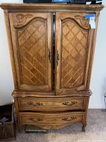 Exterior front view of wooden armoire cabinet with decorative carved door panels and metal handles on doors and drawers.