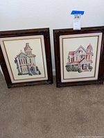 Two framed cross-stitch artworks of Victorian houses side by side on carpeted floor under wall