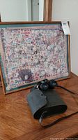 Photo showing vintage Empire binoculars with attached black leather case placed in front of framed baseball puzzle art on a table.