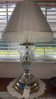 Front view of the lamp showing the glass base, brass base plate, and white fabric lamp shade with fringe.