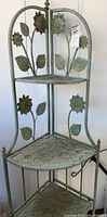Full view of metal corner rack showing three woven metal shelves and sunflower decorative pattern on the back panels.