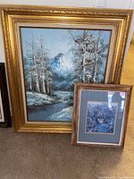 Two framed artworks side by side on carpeted floor showing a large oil painting in ornate gold frame and smaller framed print.