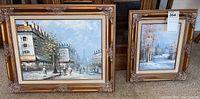Two framed oil paintings with ornate gold frames: one urban street scene and one winter forest scene.
