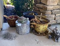 Overview of three outdoor ceramic pots, two with wooden statues topped with concrete turtles, dirt and dead leaves visible inside pots.