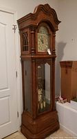 Front view of tall oak Grandfather clock showing brass dial, arched bonnet and glass front door.