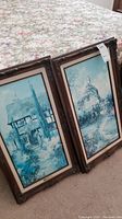 Two framed oil paintings leaning against a bed with a floral bedspread, showing blue-toned scenic houses.