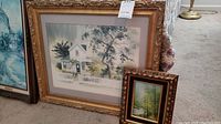 Wide view showing a large framed watercolor painting of a white house with a picket fence alongside a smaller framed oil painting of a river landscape.