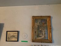 Gold ornate framed watercolor of cathedral interior alongside black framed print on wall.