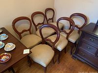 Wide shot of six Victorian style dining chairs arranged side by side on hardwood floor next to wooden furniture and dining table.