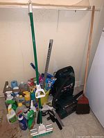 A group of various cleaning supplies including sprays, liquids in bottles, a mop with a green handle, a broom with wooden handle, and other cleaning tools arranged on floor.