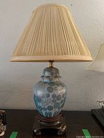 Full view of the ceramic base lamp with beige pleated lampshade, showing the entire lamp standing on a flat surface against a wall.