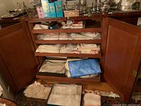 Open cabinet with three shelves and two lower drawers filled with folded linens and boxes beneath.