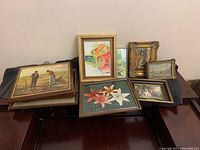 Shows several framed prints including a floral print, a print of a couple in a field, and several smaller framed prints with various subjects on a piano surface.