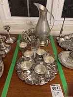 Wide view of the metal dish collection on a wooden table, showing silver tray with six coffee cups, large jug, candlesticks, and more