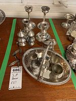 Photo showing overall collection of silver plated items arranged on a wooden surface including candlestick holders, salt and pepper shakers, napkin rings, and a metal tray with handle.