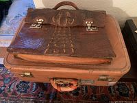 Top-down view of a vintage suitcase with an alligator skin briefcase placed on top, showing the texture and metal clasps of the briefcase.
