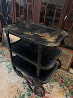 Side view showing the three black wooden shelves with intricate Asian style painted scene on the top tier and large spoked wheels on the side.