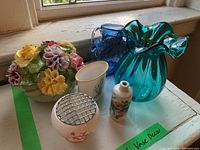 Wide shot showing six decorative vases: teal ruffled glass vase, cobalt blue glass pitcher, porcelain floral arrangement and three small porcelain vases