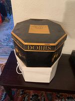 Closed octagonal Dobbs brand hat box, black and gold design, placed on top of a white box, showing the exterior details and branding of the men's hat box.