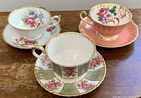 Three vintage porcelain tea cups and saucers shown from an angled top side view on a wooden surface, displaying floral patterns and gold accents.