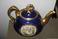 Antique KPM Meissen porcelain teapot showing cobalt blue body with gold handle, spout, and trim. Central hand-painted pastoral scene of two women in period dress. Photograph captures front view with slight angle to show lid and handle detail.