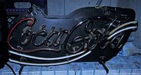 View of Coca-Cola neon sign from front showing red neon 'Coca-Cola' text and two white stripes beneath on black backing.