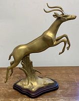 Side angle of brass leaping springbok mounted on dark wooden base, showing full body and horn detail.