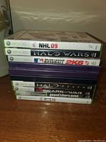 Stack of 10 Xbox 360 game cases showing titles on spine: NHL 09, Halo Wars, MLB 2K6, Halo Reach, Halo 3, Gears of War, Grand Theft Auto IV, NHL 14.