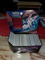 Open tin case showing a large stack of Pokémon trading cards inside, featuring Sylveon artwork on the lid.