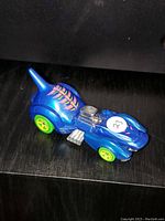 Photo of a single Hot Wheels blue toy car with shark fin, yellow wheels, and detailed engine, placed on dark surface.
