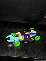 Photo shows a single Hot Wheels toy car, purple body with green wheels, blue front prongs, and yellow side details.