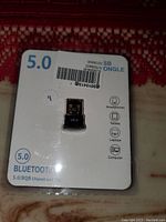 Photo of a new USB Bluetooth 5.0 dongle packaged on a white card with printed specifications and compatible device icons.