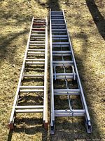Two aluminum extension ladders laid flat on the ground, showing the overall size and structure of each ladder side by side.