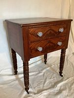 Full front and side view of the antique end table showing two drawers with clear glass knobs and turned legs with casters.