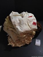 Stack of eight garments topped by folded white pullover Red Cross dress with red cross emblem and pinned notes.