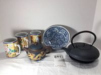 Complete set of Asian-inspired decor items displayed together: 3 hexagonal coffee mugs with colorful birds and flowers, a small phoenix and dragon themed ceramic teapot, a blue and white Andrea by Sadek bowl, and black metal teapot with trivet.
